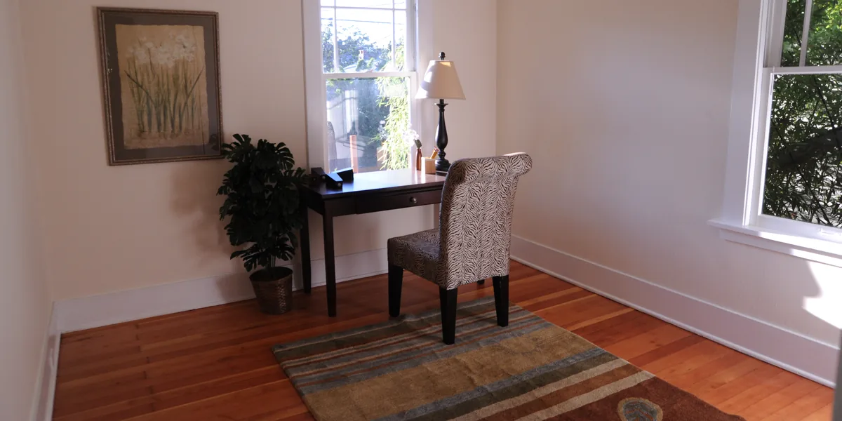 Habitación con suelo de madera: un escritorio oscuro ante una ventana, una lámpara de sobremesa de tulipa clara, una silla tapizada y una alfombra de rayas