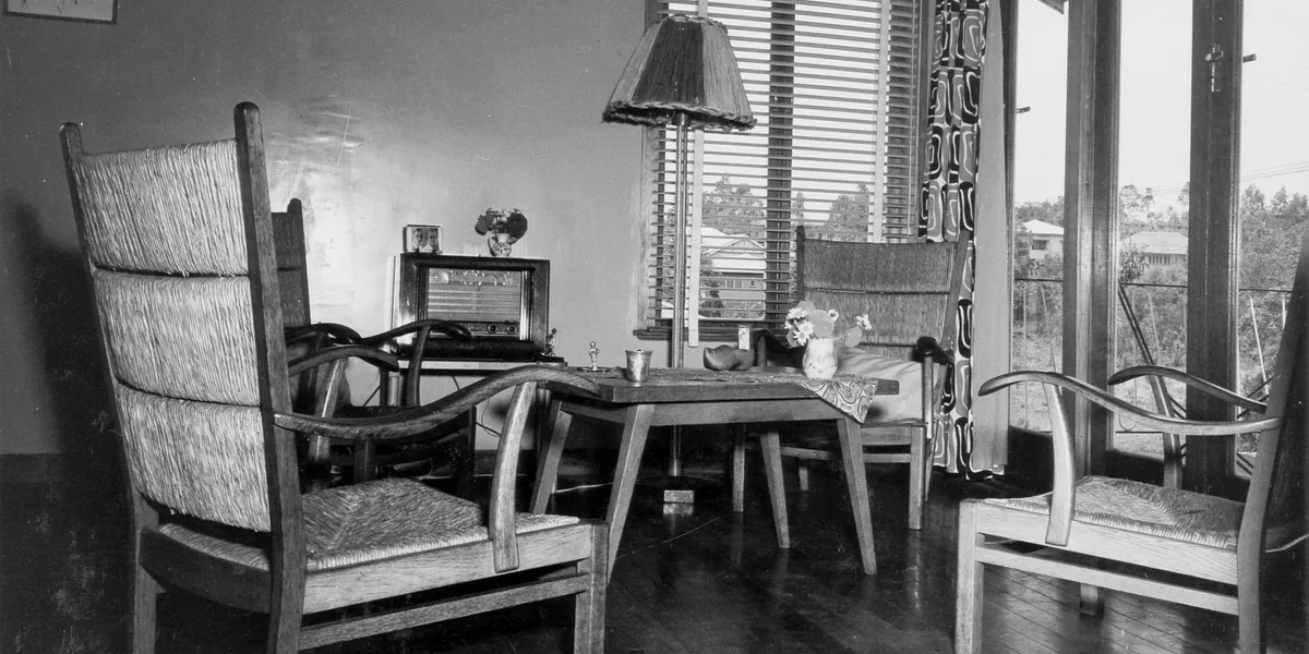 Fotografía en blanco y negro de una sala con butacas de madera y enea, una mesa baja y una lámpara de suelo con tulipa de flecos ante una ventana con persiana